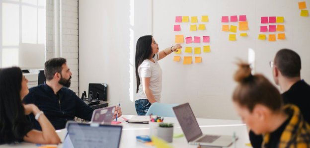 People sat around a table typing on laptops. One person is stood at the front pointing at square sticky notes stuck on a whiteboard. 