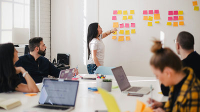 People sat around a table typing on laptops. One person is stood at the front pointing at square sticky notes stuck on a whiteboard.