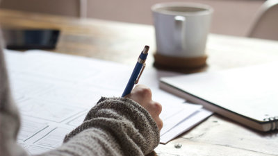 Person sat at a wooden table writing on paper near a white ceramic mug.