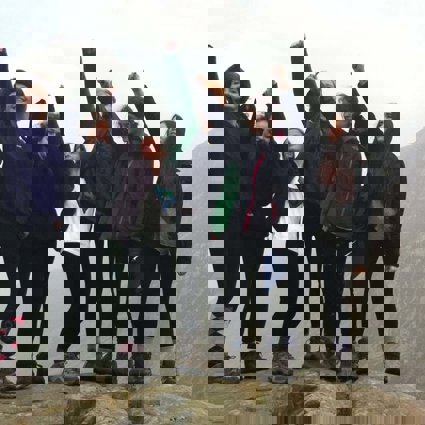 A group of young women standing on a rock in the outdoors with their arms reaching int the air, clearly enjoying geography