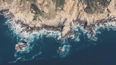 An aerial image of a rocky coastline edge, with mud and vegetation on the cliffs and waves breaking around the cliff foot.