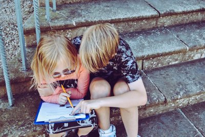Two Children sitting together on a step, one is holding a pencil and has a clipboard on their knee, the other is pointing at the clipboard with their finger