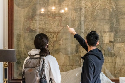 People stand in from of a large wall mounted map with their back to the camera as they look and point at the map.