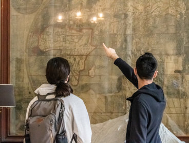 People stand in from of a large wall mounted map with their back to the camera as they look and point at the map.