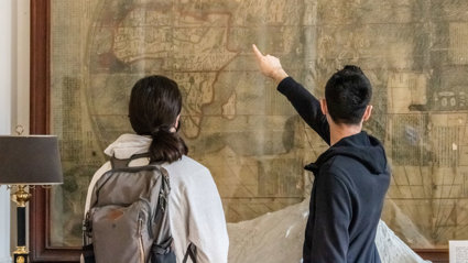 People stand in from of a large wall mounted map with their back to the camera as they look and point at the map.
