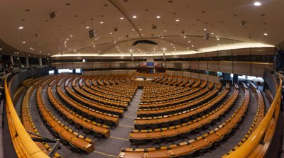 An elevated view of a very large auditorium filled with brown wood desk and black office chairs. The European Union flag is pinned up behind the podium inbetween the flags of all the EU nation drapped on flag polls.