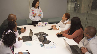 Six people sit around a white table in a board room listening to a seventh person at standing at the head of the table talk.