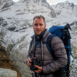 A headshot of Tom McShane in hiking gear, holding a camera on the side of a mountain.