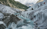 A glacial metlwater stream emerging from a glacier, which is grey streaked with black. The water is an icey blue colour. In the background are mountains