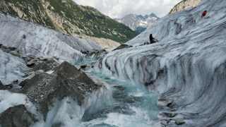 A glacial metlwater stream emerging from a glacier, which is grey streaked with black. The water is an icey blue colour. In the background are mountains