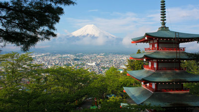 An elevated view of Tokyo, with the top of the Chureito Pagoda and trees in the foreground. The city occupies the middle ground whilst a mountain towers over it in the background.