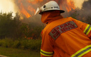 Two firemen are fighting a fire by the side of the road. Their uniforms are orange and their hose is yellow. The flames are extremally high and there is smoke