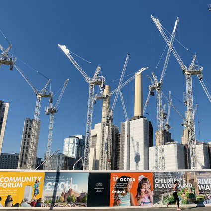 A city scene with skyscrapers and cranes to illustrate the amount of building work going on. In the foreground are colourful adverts on a boarded wooden wall around the building site.