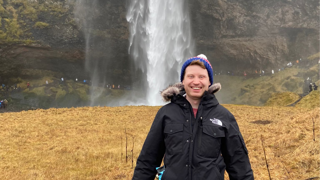 Paul Logue smiling in front of a waterfall whilst in a field with yellow coloured grass.