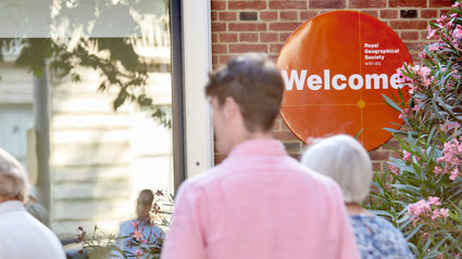 People walk into the RGS at Exhibition Road. An orange welcome sign is focused in on.