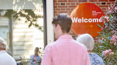 People walk into the RGS at Exhibition Road. An orange welcome sign is focused in on.