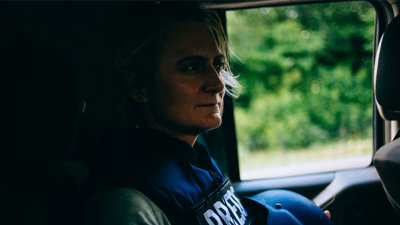 Person seated in a car wearing a vest that says 'press' and holding a protective helmet.