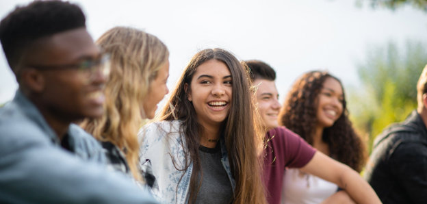 Group of five young people sitting outdoorsk, with the focus is on a brown-haired person in the centre.