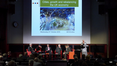 A speaker stands by a podium whilst six planalists sit in bright orange chairs, upon a dimly lit stage. a presentaion is projected on the board behind them. The title says, Cities, growth and rebalancing the UK economy. and audince can be seen in the forground watch watching. 