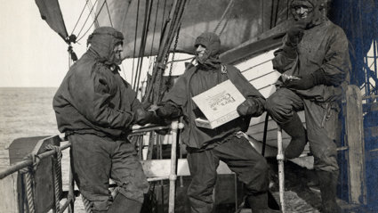 Black and white photograph of three people on a boat dressed in waterproof gear. They're smiling and eating chocolate from a box of Fry's Chocolate.