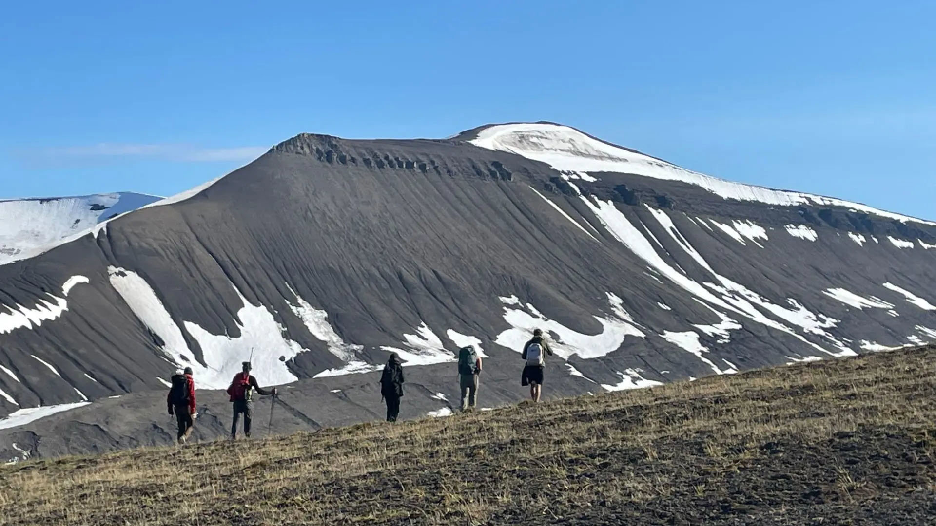Five people walking in a line through a mountainous landscape. The mountain slope is largely bare with patches of snow remaining.