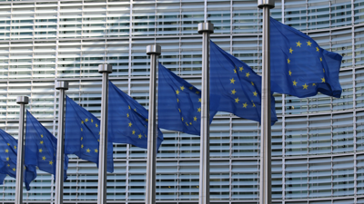 Seven European union flags each depicting a blue backgroundwith yellow stars forming a circle blow in the wind whilst on flag polls. Behind them is the chiefly metal and glass European Commission Berlaymont building.