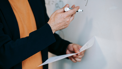 A teacher stands next to and points at a whiteboard whilst holding a whiteboard marker and paper in the other hand. There is non-distinct writing on the paper and blurred out numbers written on the white board.