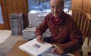 A man sitting in front of a pile of books he has published and signing one book.