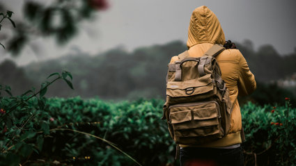 Person in coat and with backpack in a natural setting seen from behind and holding a DSLR camera.