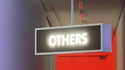 Illuminated rectangular sign with the word 'OTHERS' hanging from a ceiling in a dimly lit corridor with red and purple lighting.