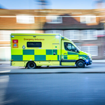 An ambulance driving at speed. The background of the picture, which is a residential street, is blurry to indicate the speed at which the vehicle is travelling.