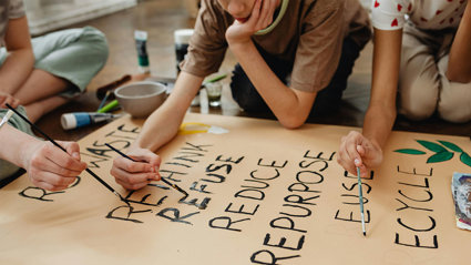 Three students painting the words rethink, refuse, reduce, repurpose, and recycle on brown paper with black paint.