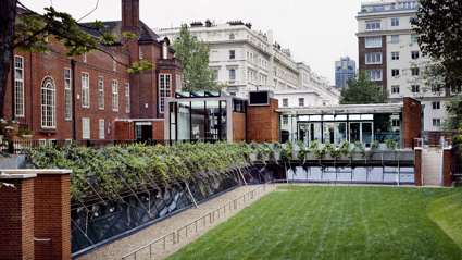 A grass covered patio bordered by a rectangular stone path. This lays surrounded by the red bricked Society building.
