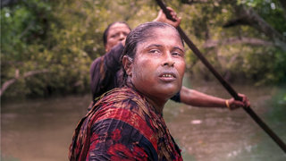 Person with wet hair in a small boat glances over shoulder while another person in the back paddles through the mangrove-lined river.