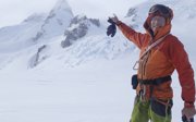 A expeditioner in full winter gear on a snow covered glacier pointing to the top of a mountain.