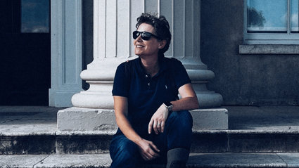 A person in dark clothes and sunglasses sits smiling on stone steps in front of a large white columned building.