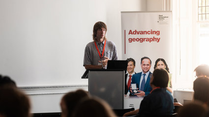 A speaker is presenting behind a lecturn. A poster next to him reads 'advancing geography'.