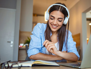Person wearing headphones smiles while studying with a notebook and laptop.