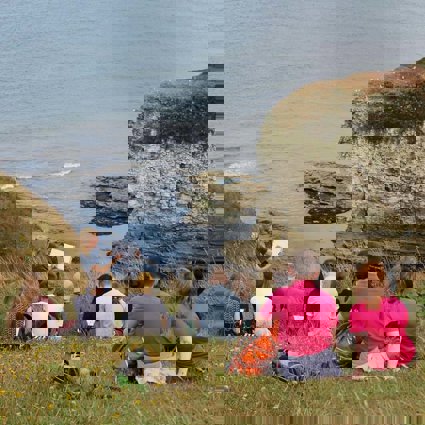 A teacher is standing at the top of a cliff, overlooking a rocky beach cove, holding a clipboard and showing a group of students something