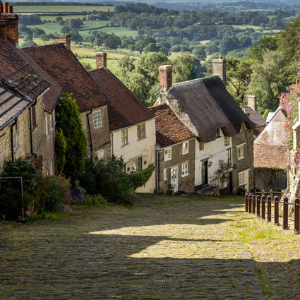 A steep village road with a cobbled roadway, a pretty sceneic village idyll. Countryside stretches into the background beyond the village