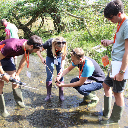 A group of teachers are standing in a river, all wearing wellies, measuring the depth of the river using a stick.