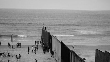 People gathered along a tall border fence extending into the ocean on a sandy beach under a cloudy sky.