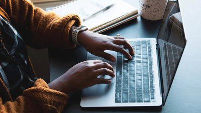A person at a desk typing on a laptop with a notebook and mug to one side.