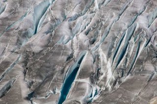 An aerial view of a crevasse field, showing grey tinted glacier ice with bright blue crevasses streaked through it