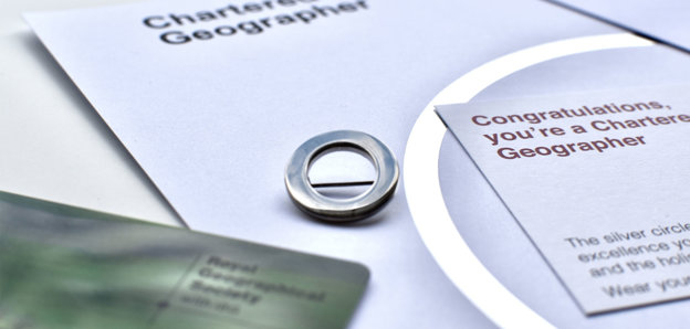 A silver circle lapel pin, a symbol of Chartered Geographer status, next to a card that says 'Congratulations, you're a Chartered Geographer'. 