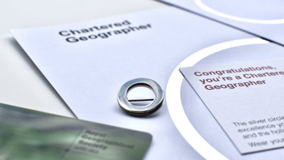 A silver circle lapel pin, a symbol of Chartered Geographer status, next to a card that says 'Congratulations, you're a Chartered Geographer'. 
