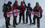 Group of people in winter gear holding up flags of Georgia and the United Kingdom while standing on top of a snow covered mountain.