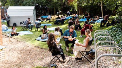 People sitting outside on a patch of grass, eating while seated on chairs and mats