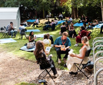 People sitting outside on a patch of grass, eating while seated on chairs and mats