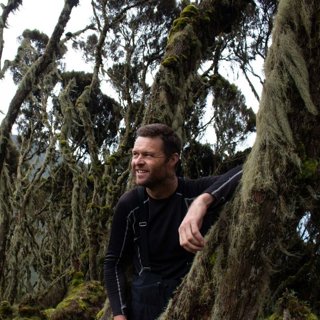 Jon Beardmore in a black long sleeved top, leaning against a mossy tree.
